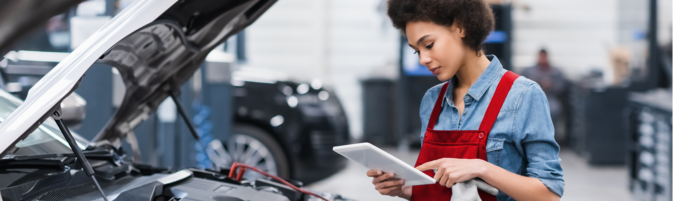 A female mechanic wearing a red apron running diagnostics on a car s engine. With a focused precision, she carefully analyzes the readings from her tablet.