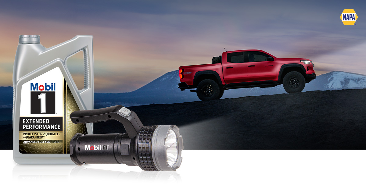 A red truck is driving up a mountain incline at dusk. A Mobil 1 motor oil bottle and illuminated flashlight are in the foreground.