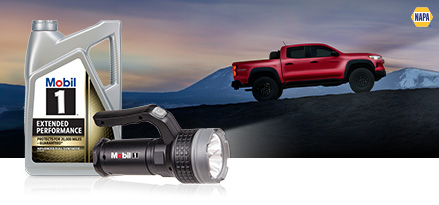A red truck is driving up a mountain incline at dusk. A Mobil 1 motor oil bottle and illuminated flashlight are in the foreground.