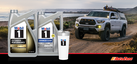 A pickup truck driving on a dirt road with water in the background. Two Mobil 1 bottles and a tumbler sit in the foreground.