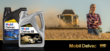 The image features two containers of Mobil Delvac motor oil prominently in the foreground, labeled "1300 Super" and "Delvac 1 ESP." Behind them, a person stands with arms crossed in a wheat field, wearing jeans and a plaid shirt. In the background, a large agricultural machine, possibly a combine harvester, is working through the field, creating a cloud of dust. The Mobil Delvac logo appears at the bottom right, along with a "100 years" emblem, indicating a centennial celebration. The overall scene emphasizes durability and reliability for heavy-duty vehicles and agricultural equipment.