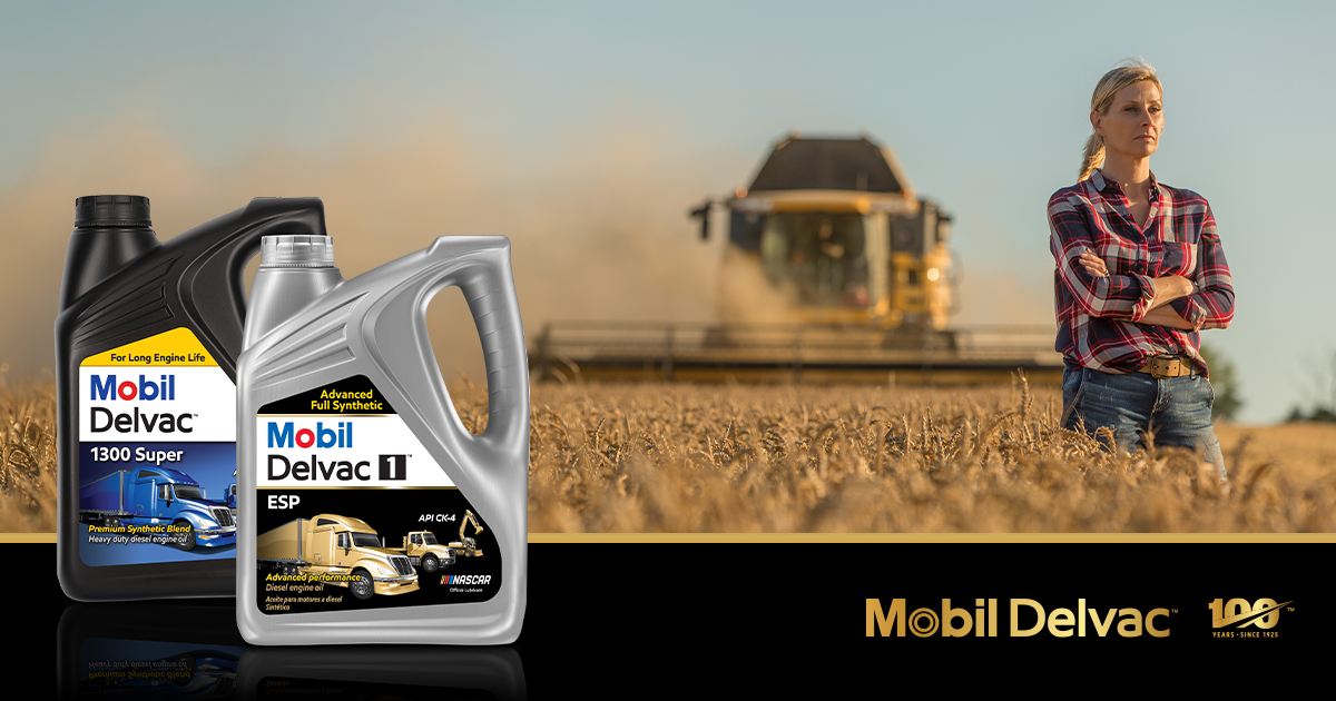 The image features two large containers of Mobil Delvac engine oil in the foreground: one labeled "Mobil Delvac 1300 Super" and the other "Mobil Delvac 1 ESP." In the background, there is a golden wheat field with a combine harvester in action, kicking up dust as it harvests the crop. To the right, a person in a plaid shirt and jeans stands confidently with arms crossed, suggesting a farming or agricultural setting. The bottom of the image includes the Mobil Delvac logo and a gold emblem marking "100 Years – Since 1925." The overall scene highlights reliability and performance for heavy-duty vehicles and equipment in agricultural work.