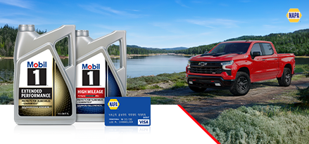 A red Chevy truck drives on a dirt and gravel road alongside a river, with lush evergreen trees lining both sides and extending all the way to the horizon.