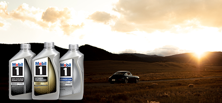 The image shows three bottles of Mobil 1 motor oil prominently displayed in the foreground. In the background, a car is parked on a rural road with rolling hills and mountains under a partly cloudy sky