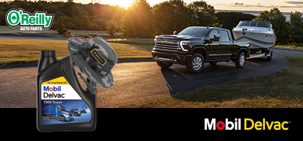 A black pickup truck is towing a white speedboat along a paved road at sunset, with trees and a lake in the background. In the foreground, there’s a large bottle of Mobil Delvac 1300 Super motor oil with a camo hat resting on top. The O’Reilly Auto Parts logo appears in the upper left corner, and the Mobil Delvac logo is along the bottom right. The scene conveys an outdoor, weekend adventure vibe.