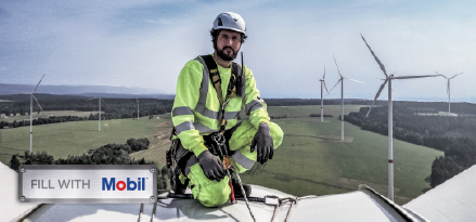 Worker in safety gear on a wind turbine overlooking a wind farm, with a Mobil sign in the foreground.