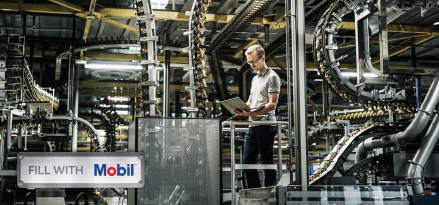Person using a tablet while inspecting automated manufacturing equipment inside an industrial production facility. The Mobil logo appears in the foreground.