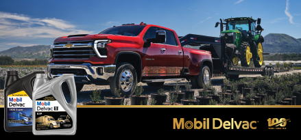 A bright red heavy-duty Chevrolet pickup truck is parked on a gravel lot, towing a flatbed trailer carrying a green tractor. Rows of small potted plants line the foreground, with mountains and a blue sky in the background. In the lower left, two containers of Mobil Delvac motor oil are displayed prominently. A black banner along the bottom features the Mobil Delvac logo and a “100 years” mark.
